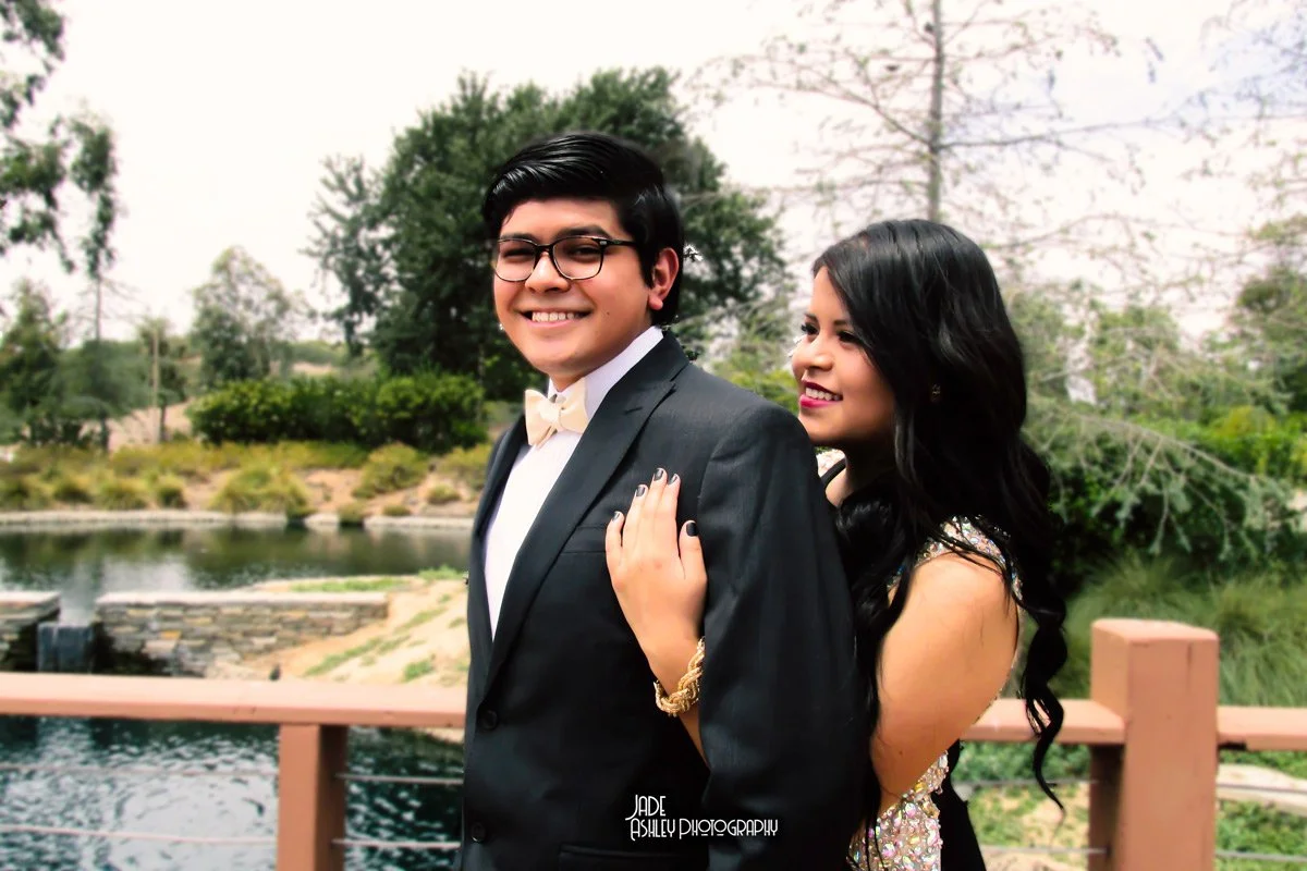 A young man in a tuxedo and bow tie and a young woman in a fancy dress pose outdoors near a pond, smiling, with trees and a bridge in the background.