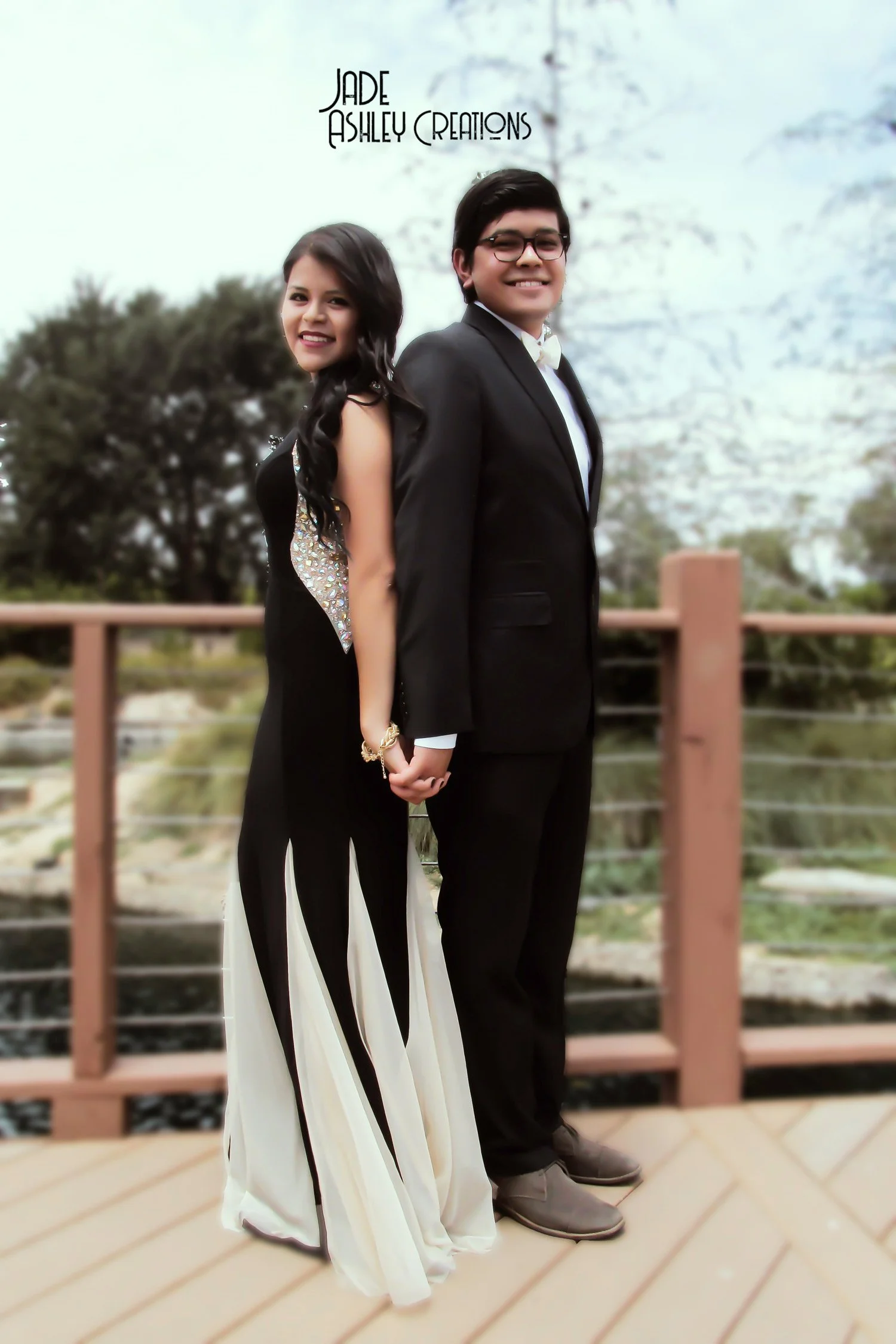 A young couple dressed in formal attire standing and holding hands on a wooden deck outdoors, with trees in the background.