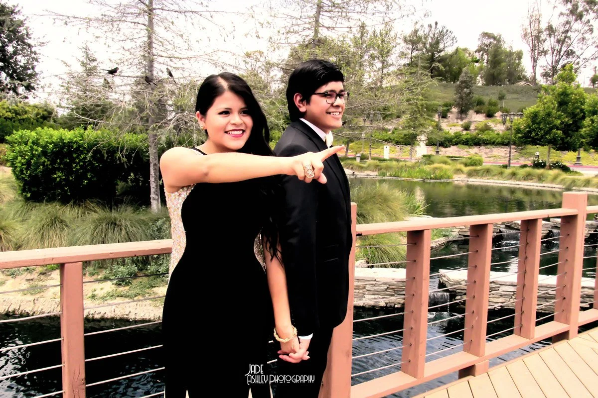 Two young adults, a woman and a man, stand on a wooden bridge by a pond in a park. The woman, with black hair and a black dress, points ahead with her right arm extended. The man, with glasses and dark hair, is dressed in a suit and smiles while look