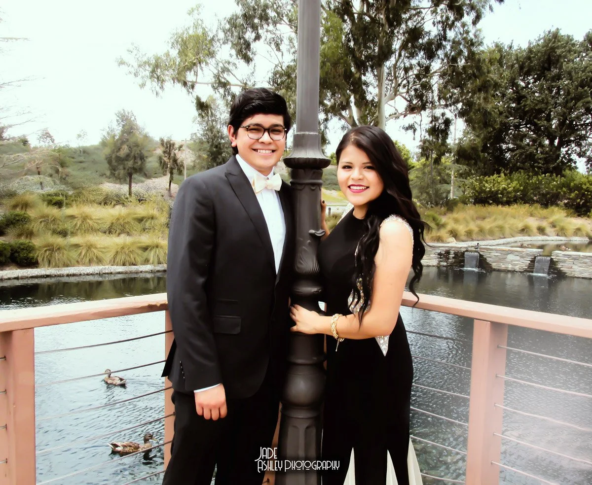 A young man and woman standing outdoors near a lake, smiling and dressed in formal attire, with trees in the background.