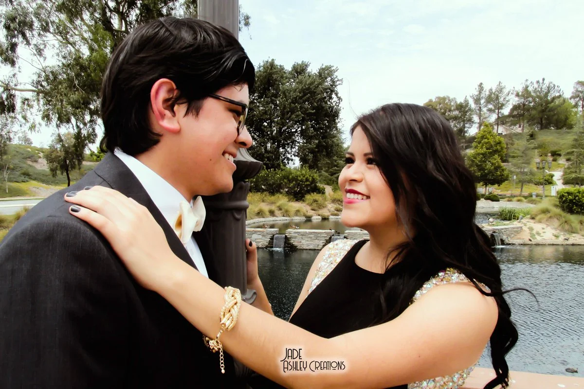 A couple dressed in formal attire standing close together outdoors by a pond, smiling at each other, with trees and a small waterfall in the background.