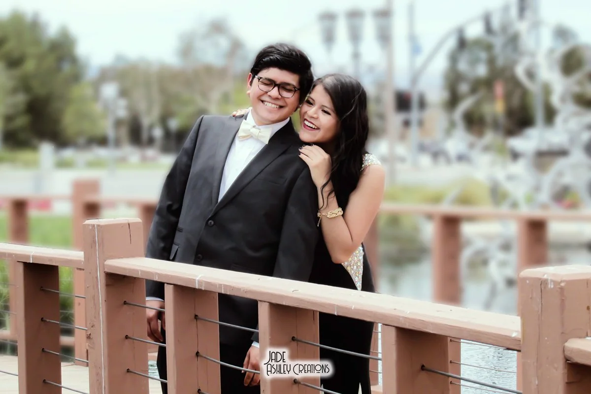 A young couple dressed in formal attire, smiling and sharing a happy moment on a wooden bridge outdoors.