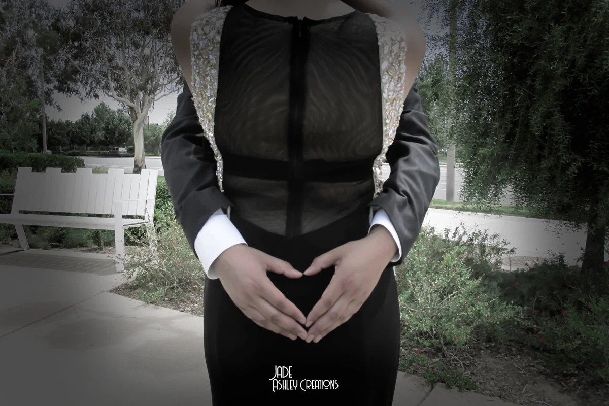 A woman wearing a black dress with sheer top and embellished shoulders, making a heart shape with her hands in an outdoor park setting.