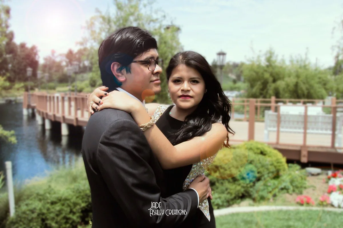 A young man and woman embrace outdoors near a pond with a wooden bridge and green trees in the background, on a sunny day.