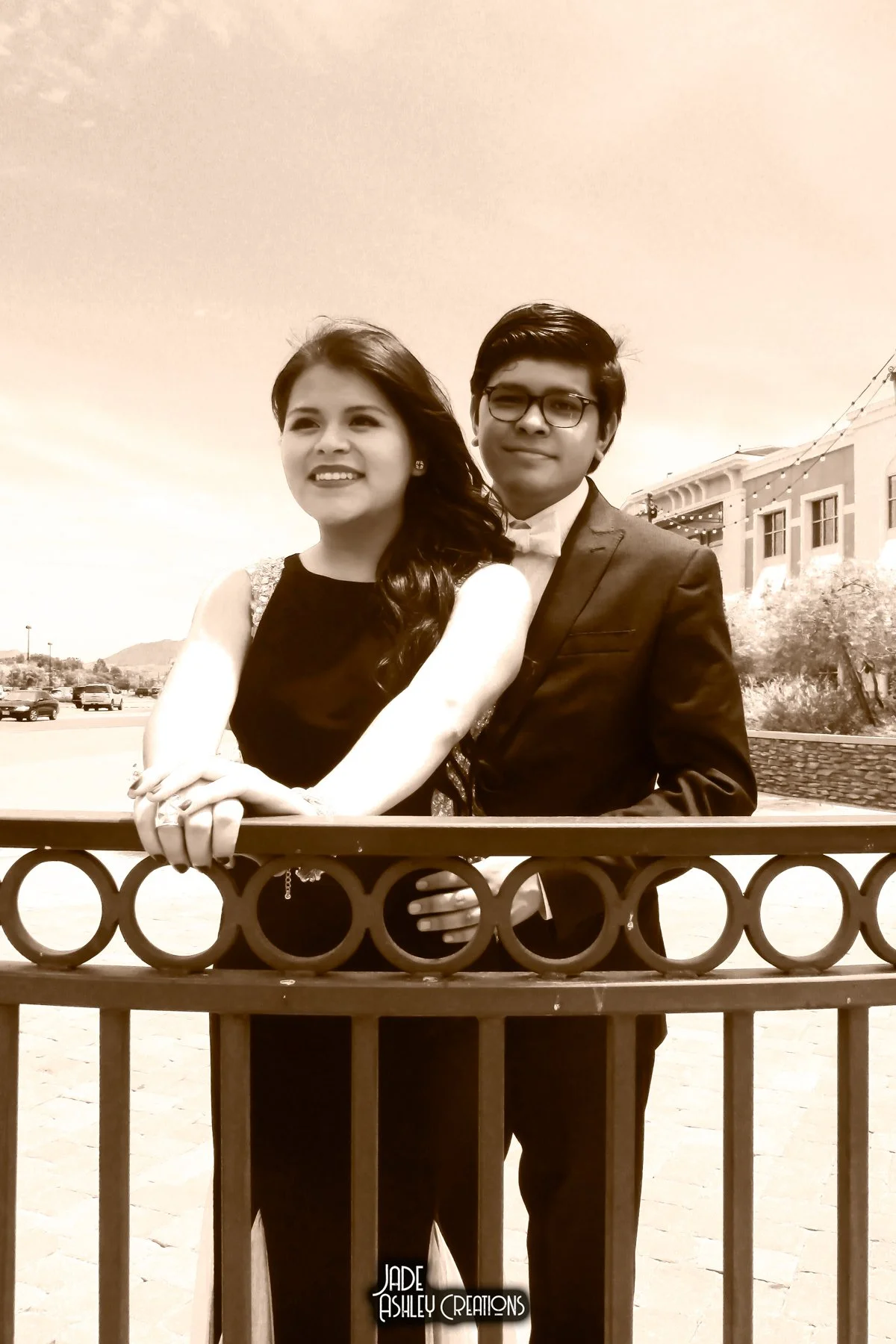 A young woman and a young man dressed in formal attire, standing behind a decorative fence outdoors, smiling. The woman has long dark hair, and the man is wearing glasses. The photo has a sepia tone.