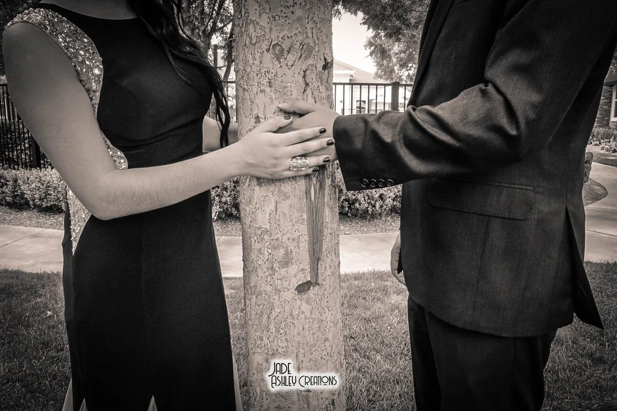 A woman and man holding hands around a tree, with the woman's hand resting on the man's arm. The woman is wearing a dress with sequin details on the sleeves, and the man is wearing a suit. They are outdoors with a fence, bushes, and homes in the back