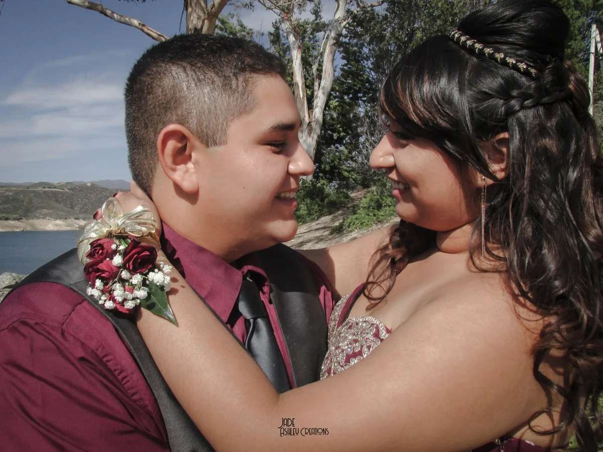 A young man and woman are smiling and embracing each other outdoors near a body of water and trees, with clear skies.