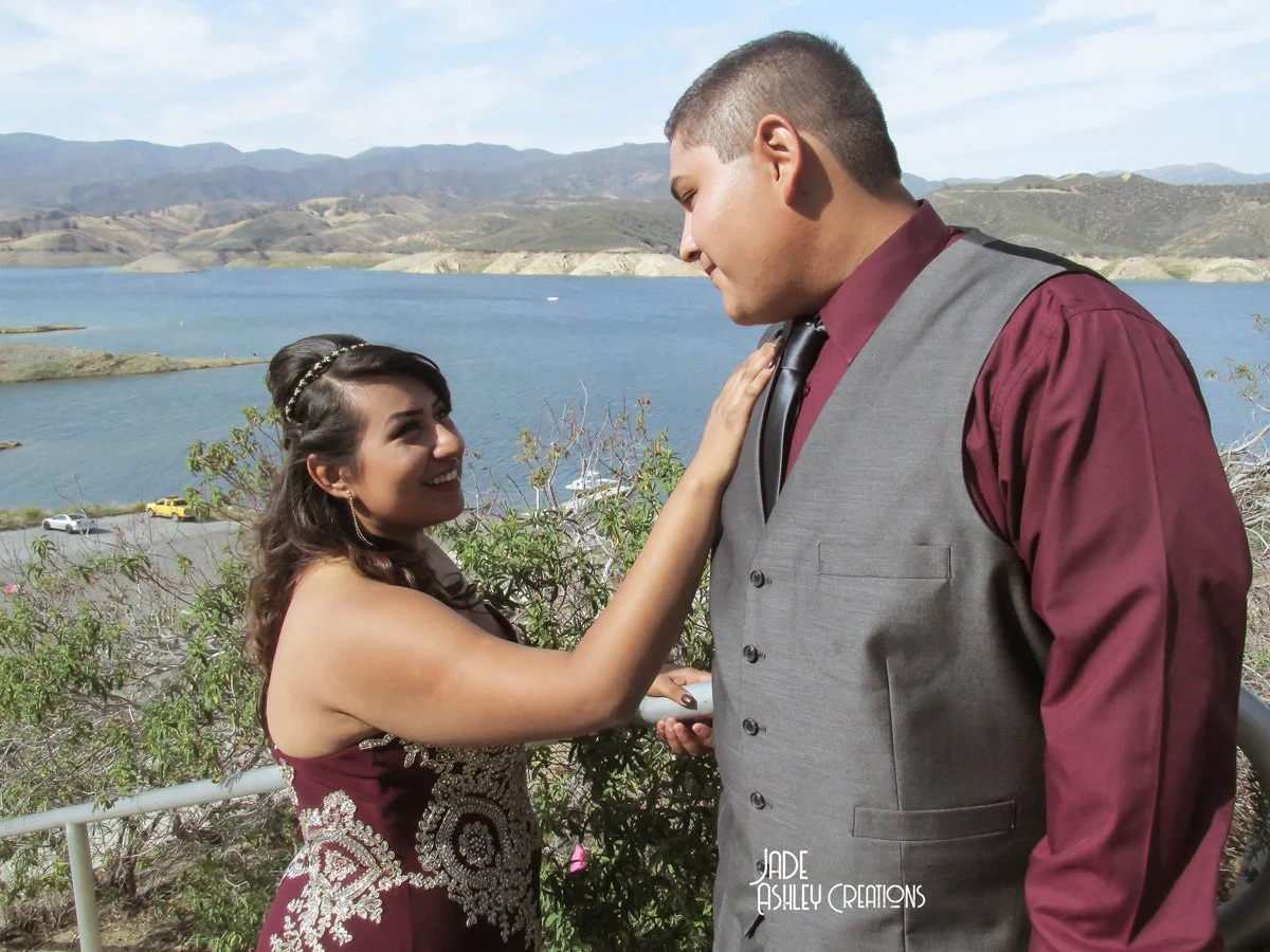 A young couple looks at each other with the woman touching the man's chest outdoors near a lake with mountains in the background.