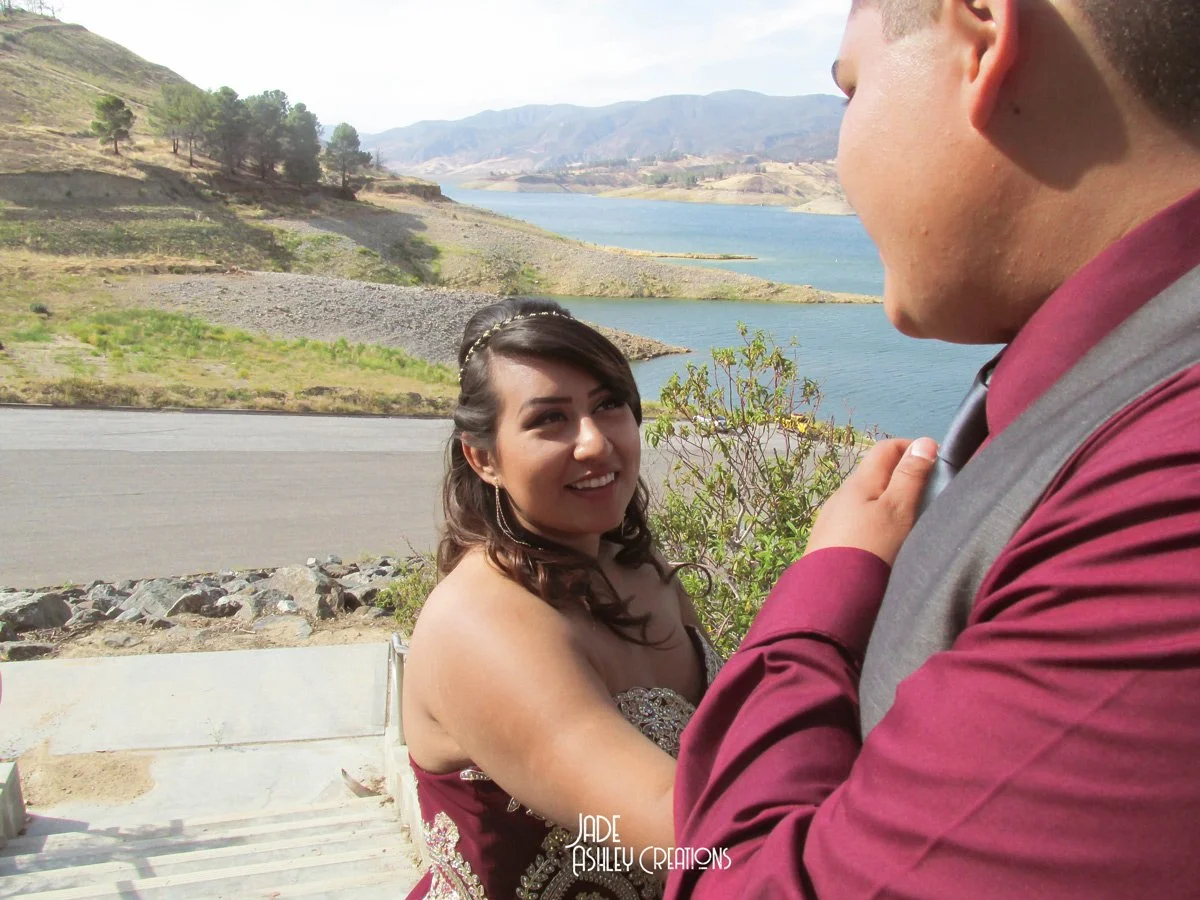 A couple standing outdoors near a body of water and mountains, with the woman smiling at the man who is dressed in a maroon shirt and gray vest.