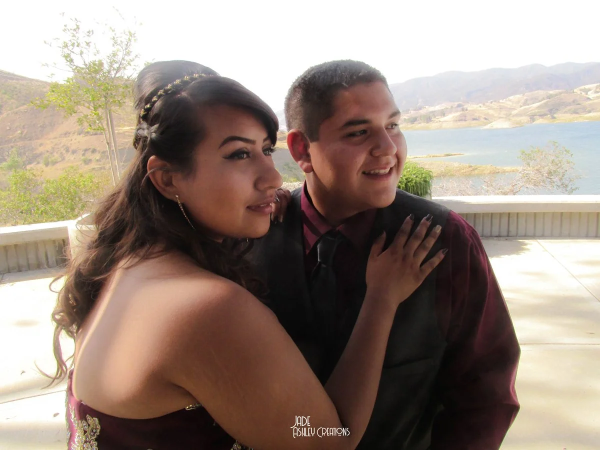 A young woman with dark hair and a young man with short hair standing closely together outdoors with a lake and mountains in the background, both smiling and appearing happy.