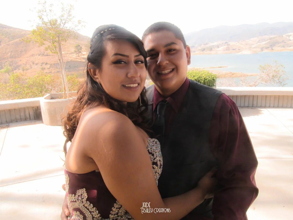 A young couple dressed in formal attire hugging outdoors with a scenic lake and mountains in the background.