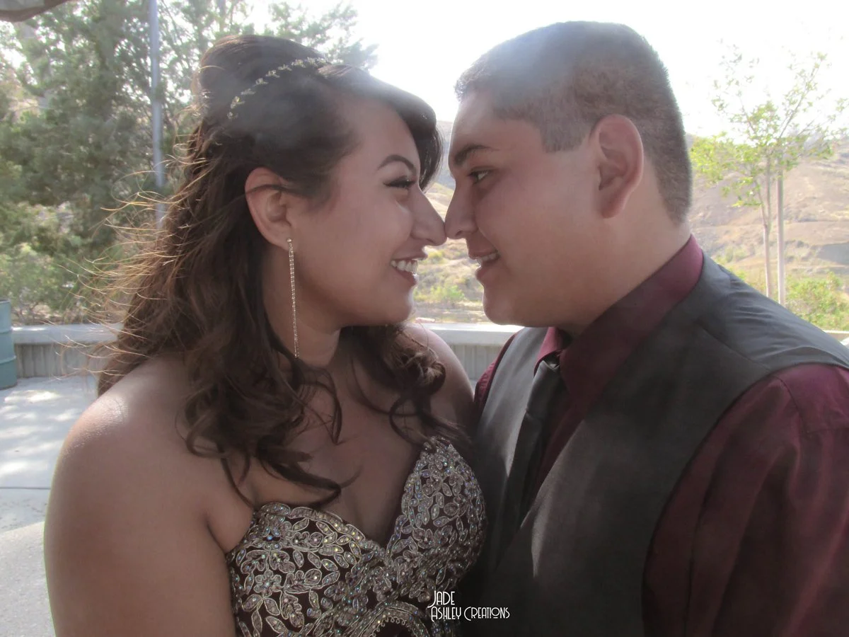 A couple is face to face outside, smiling and about to kiss. The woman has long, brown, curly hair and is wearing a strapless, ornate dress with sparkling details, and a long earring. The man has short brown hair, is dressed in a maroon shirt and a d