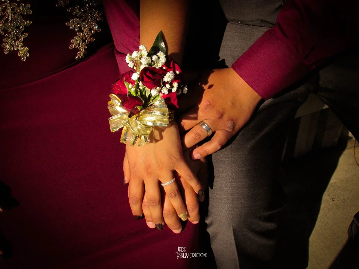 Couple holding hands, with a corsage of red roses and white baby's breath, wearing rings, dressed in formal attire.