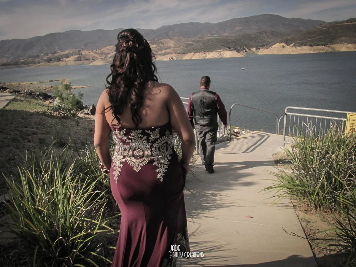 A woman in a strapless, embroidered gown walks along a lakeside path, with a man in a vest also walking ahead, overlooking a large body of water with mountains in the background.