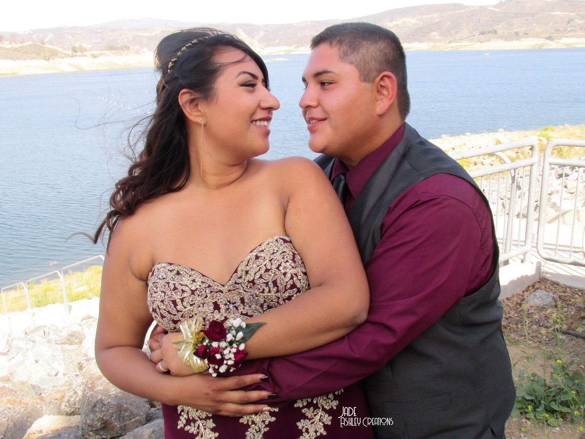 A couple dressed in formal attire embraces near a body of water with a landscape of hills in the background.