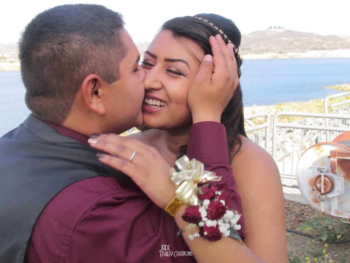 A couple about to kiss outdoors near a body of water, with a railing and landscape in the background.