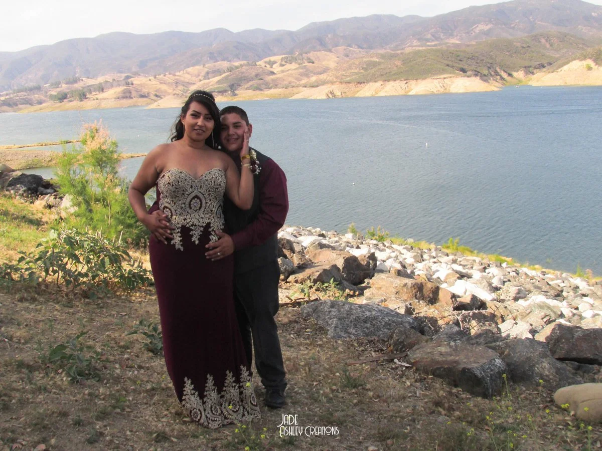 A young couple poses by a lake with mountains in the background, dressed formally, possibly for a special occasion.