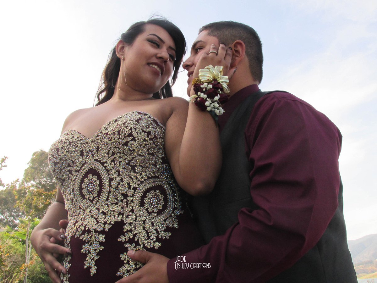 A couple dressed in formal attire, with the woman wearing a beaded, strapless dress and the man in a maroon shirt and black vest, holding each other closely outdoors during the daytime.