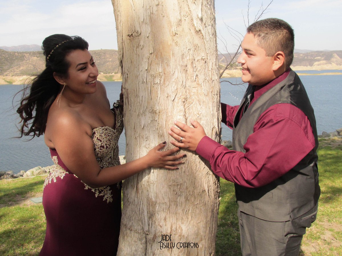 A woman in a dark purple strapless dress and a man in a gray vest and maroon shirt are touching opposite sides of a tree, smiling at each other near a body of water with hills in the background.
