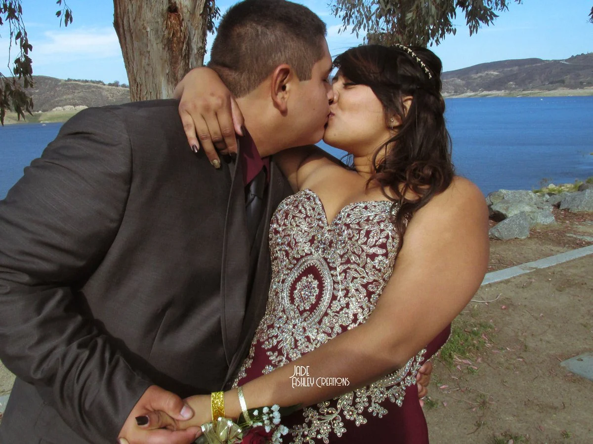 A couple kissing outdoors near a lake, with trees and hills in the background.
