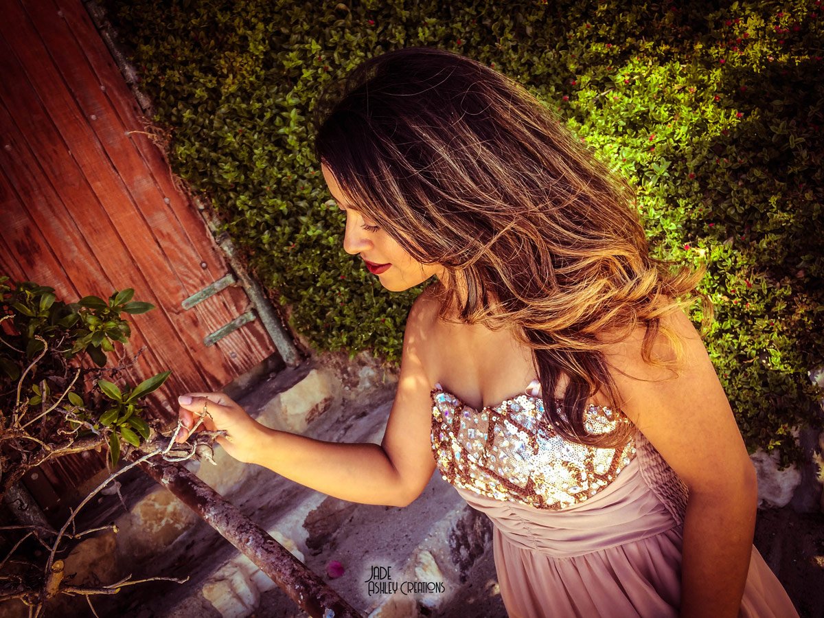 Woman with shoulder-length wavy hair wearing a sequined strapless dress, touching a small tree or bush, standing outdoors near a wooden fence and green shrubbery.