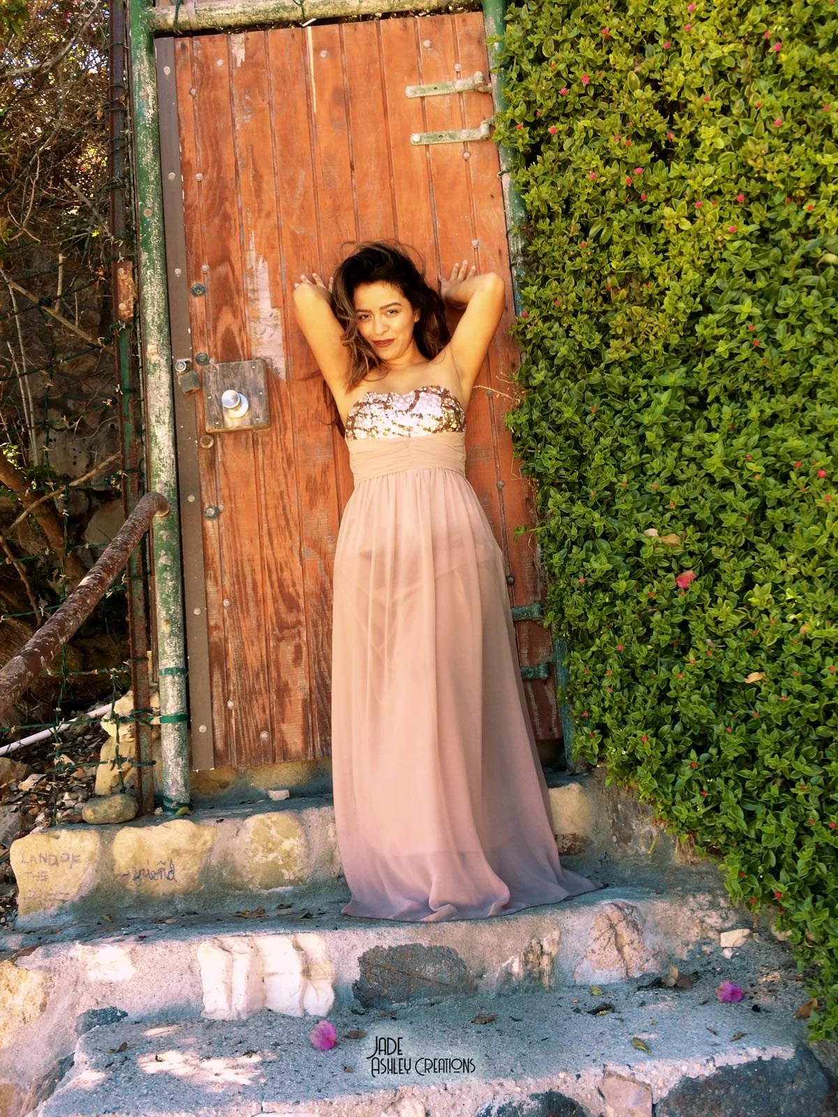 A woman wearing a strapless beige gown with a sequin top, standing on stone steps in front of a wooden gate and green foliage, with her arms raised above her head.