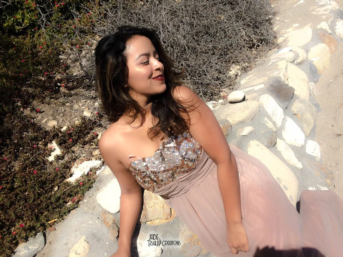 Woman in a strapless dress with sequined bodice and flowing skirt, sitting on rocks near a desert landscape with shrubs.
