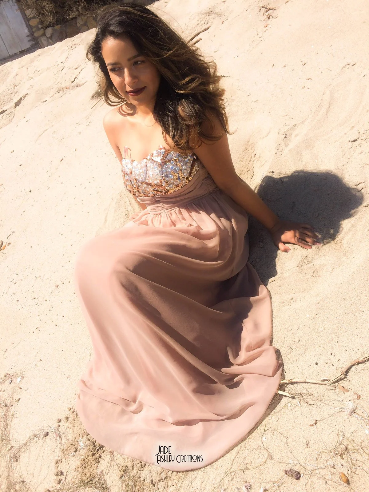 Woman in a strapless, sequined top and long beige skirt sitting on sandy ground in sunlight.