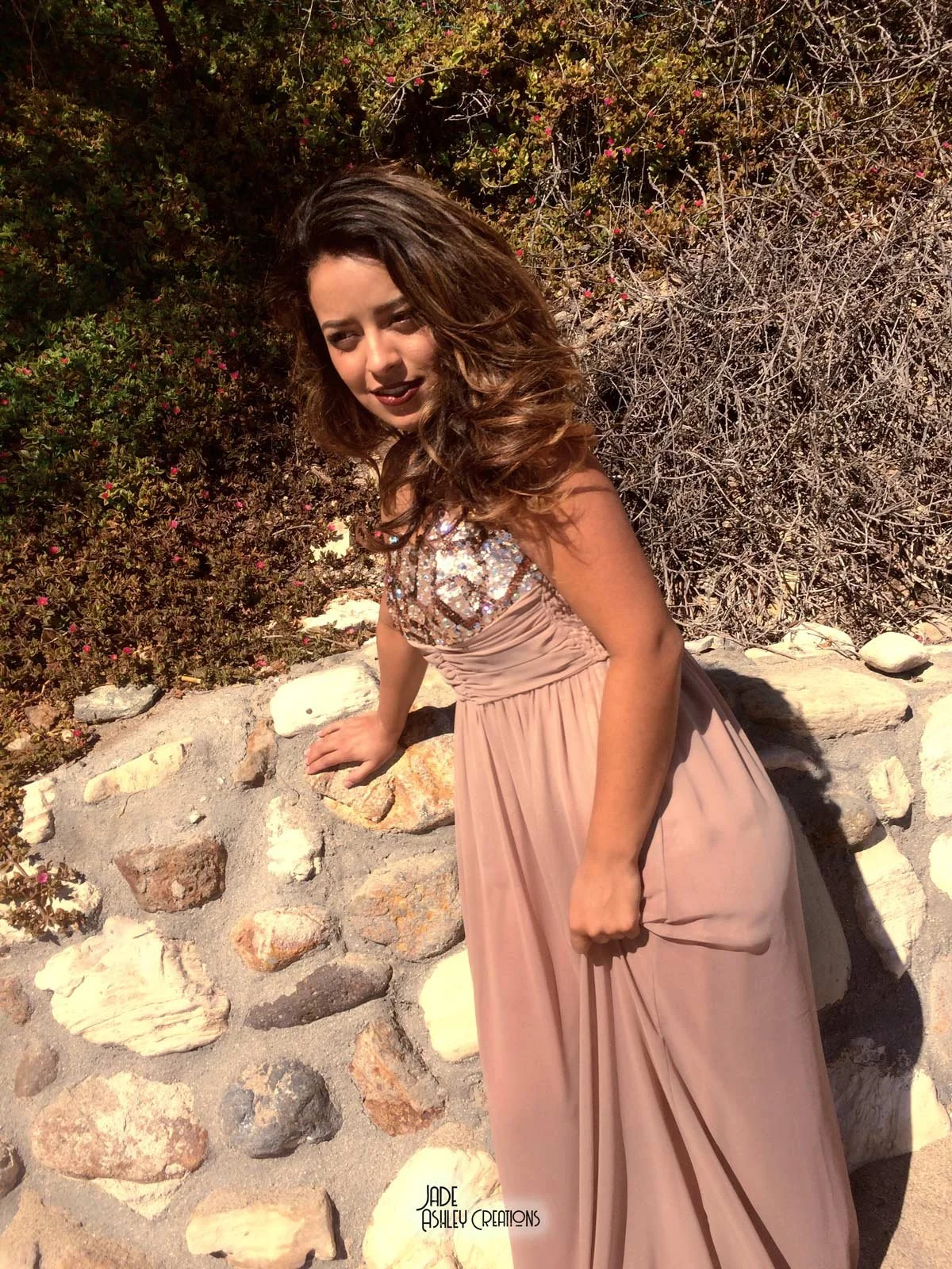 A woman in a light pink gown with sequin embellishments on the bodice, standing outdoors next to a stone wall and bushes, sunset lighting.