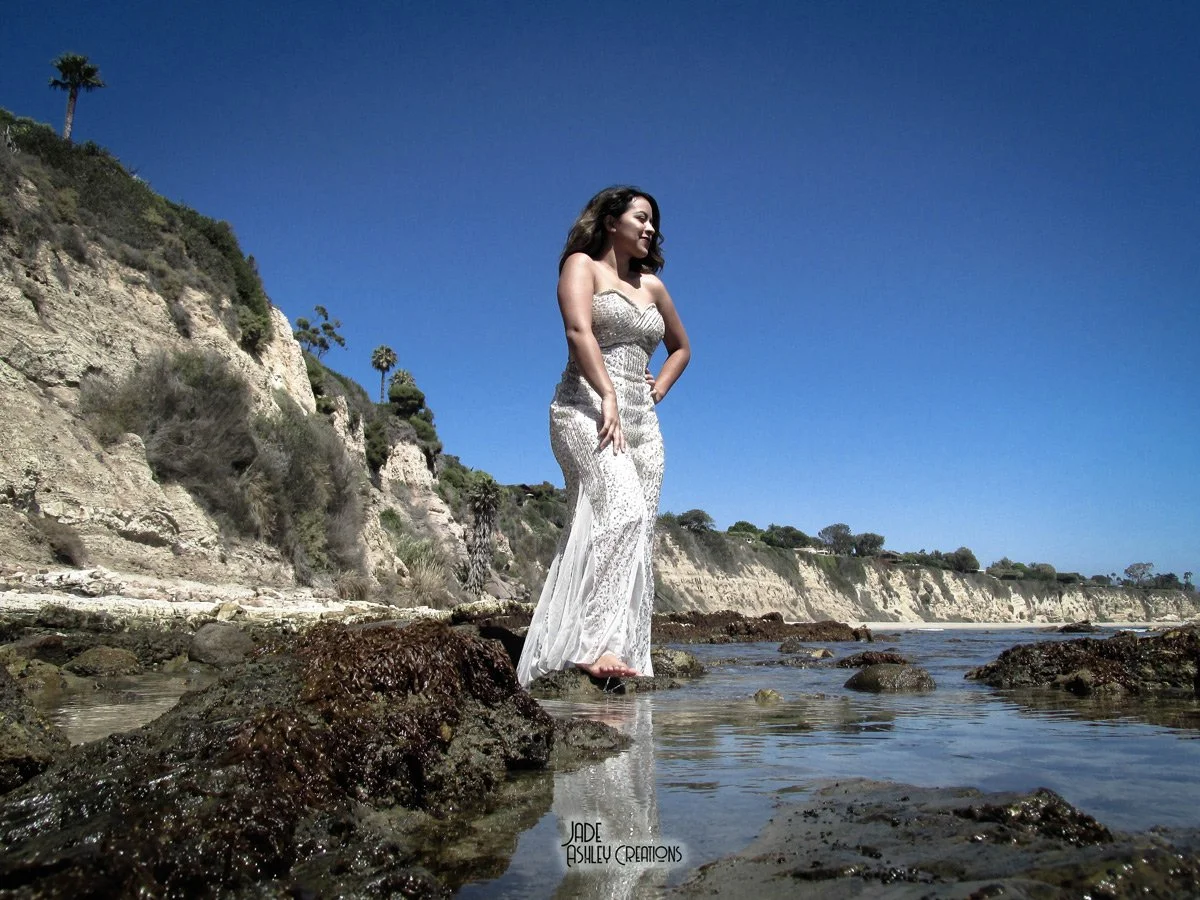 A woman in a white dress standing on rocks at the beach with cliffs and trees in the background under a blue sky.