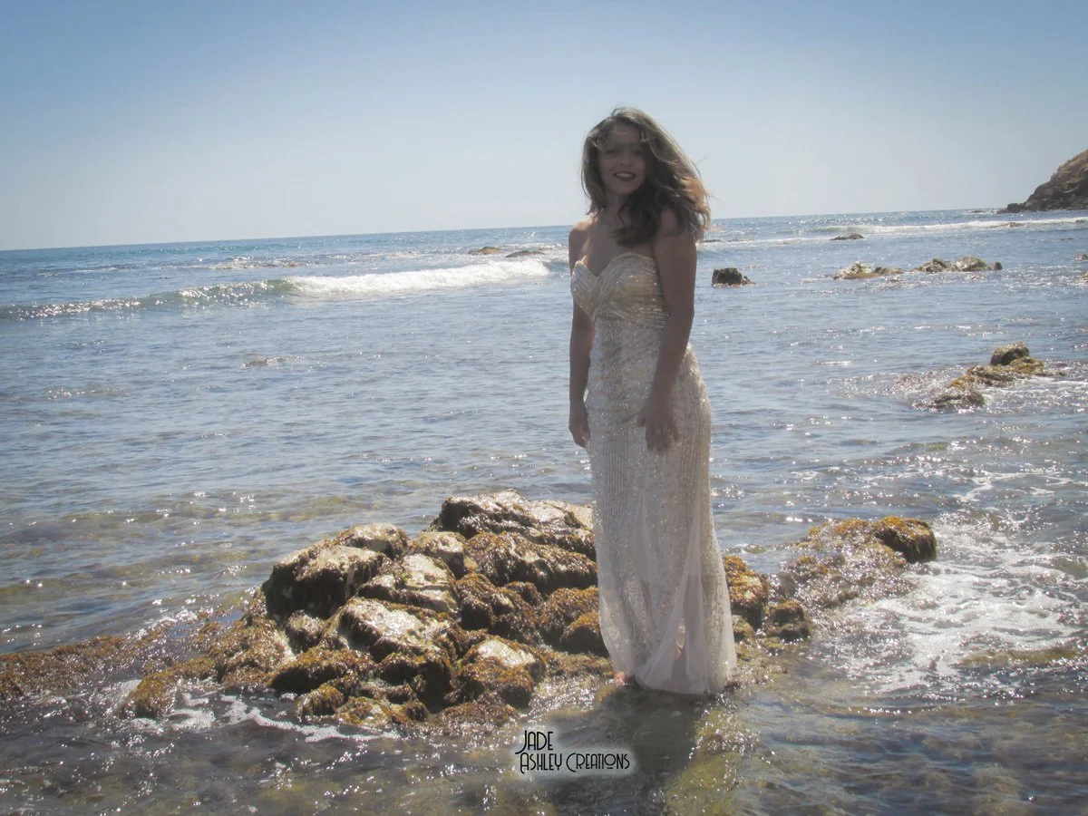 A woman in a white strapless dress stands on rocks at the edge of the ocean, smiling, with waves in the background, under a clear blue sky.