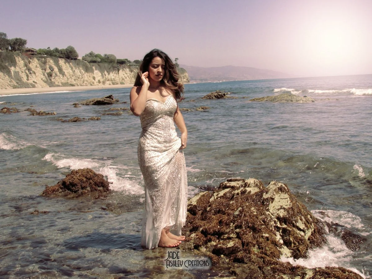 A woman in a sparkly, white, strapless dress standing barefoot on rocks in the ocean during daytime with cliffs and a coastline in the background.