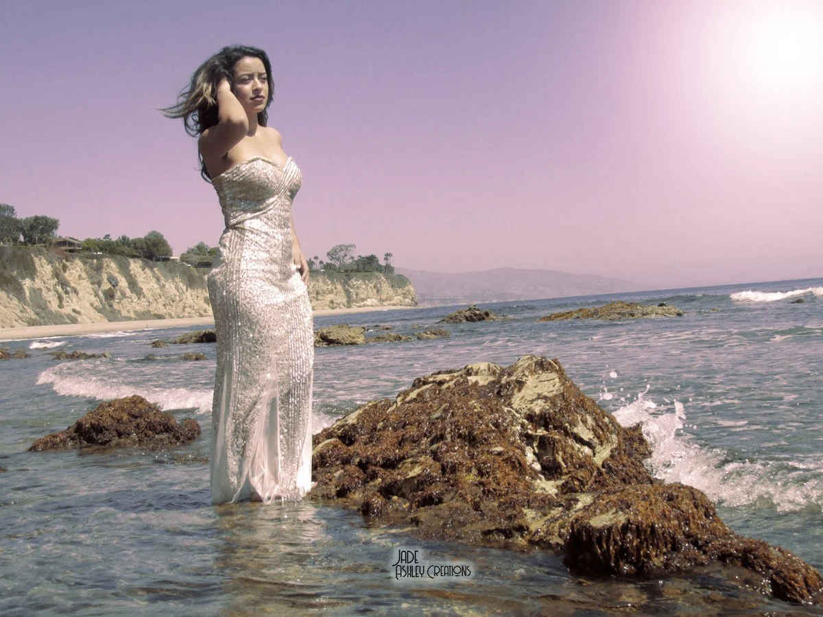 A woman in a shimmering, strapless gown stands on rocks in shallow ocean water near a beach with cliffs and trees in the background during daytime.