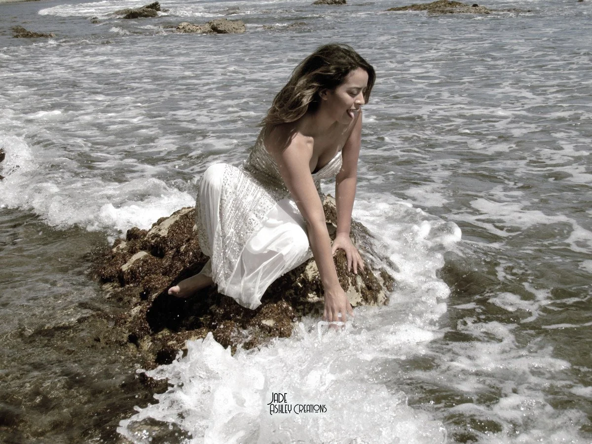 A woman wearing a white dress sitting on a rock at the shoreline with ocean waves around her.