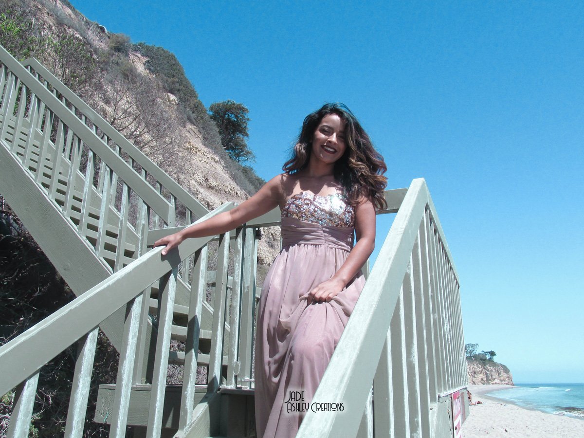 A woman in a strapless dress standing on a staircase by the beach with cliffs and blue sky in the background.