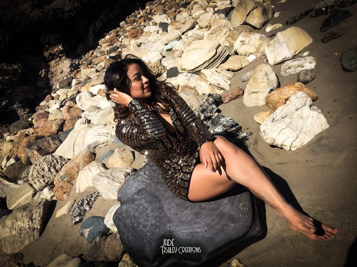 A woman in a sequined dress with black and gold tones reclining on a large gray rock at a beach surrounded by rocks and sand.