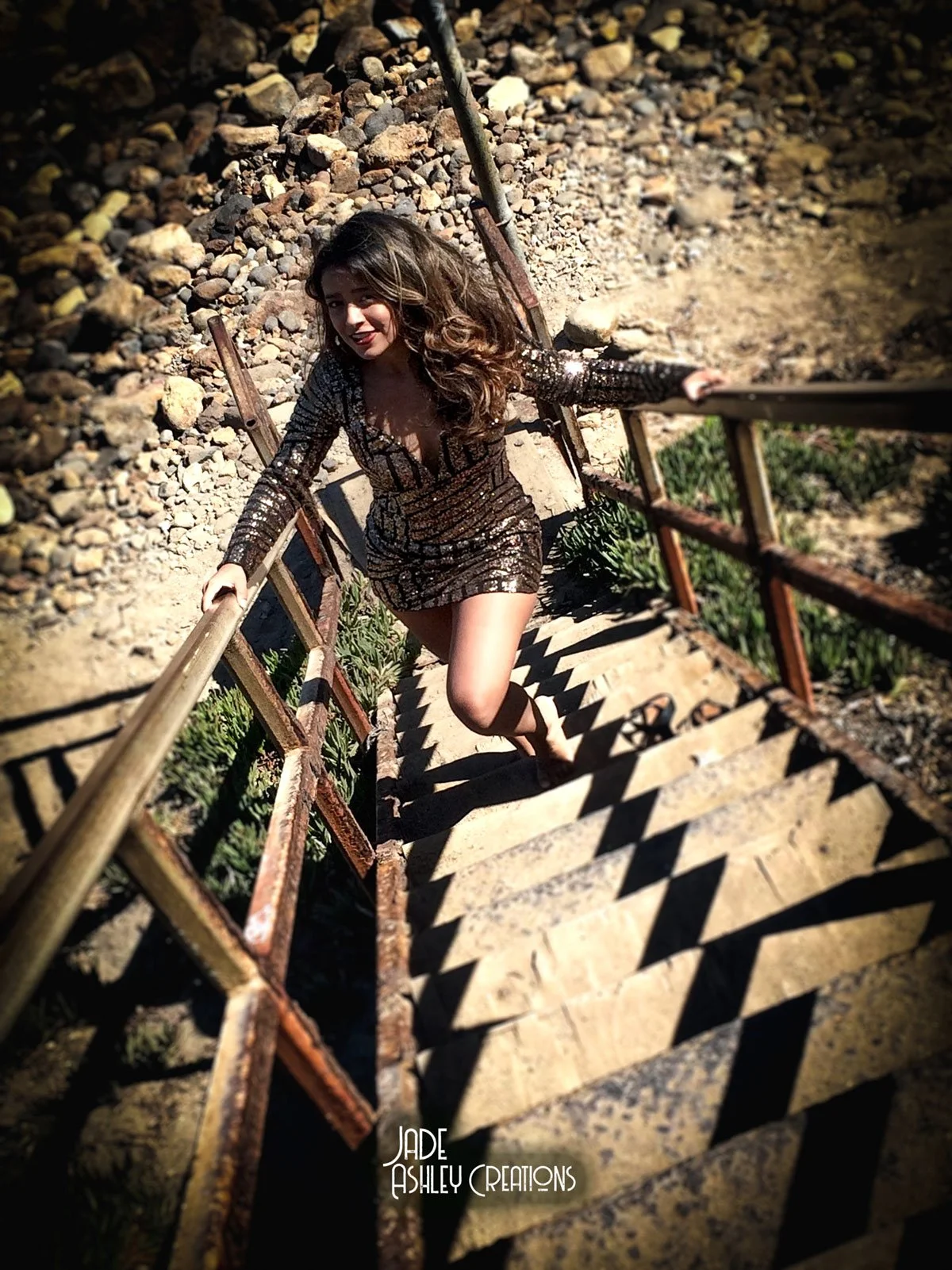 Woman in a sequined dress climbing a rusty staircase outdoors, shadowed by sunlight.