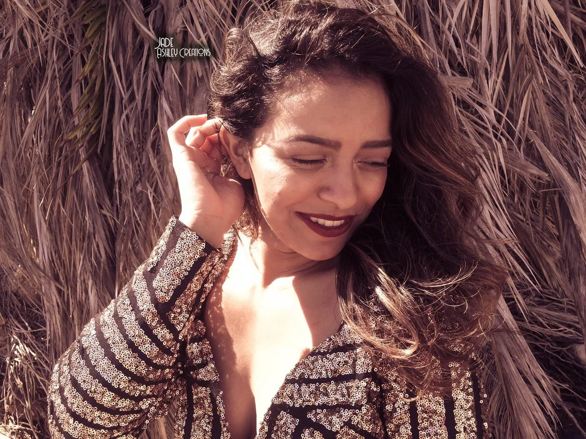 A woman with long, wavy brown hair is smiling with her eyes closed, standing in front of tall, dry grass. She is wearing a sequined top.