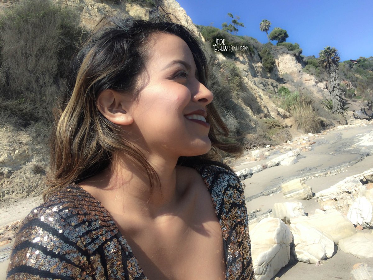 A woman smiling on a beach with rocky cliffs and palm trees in the background.