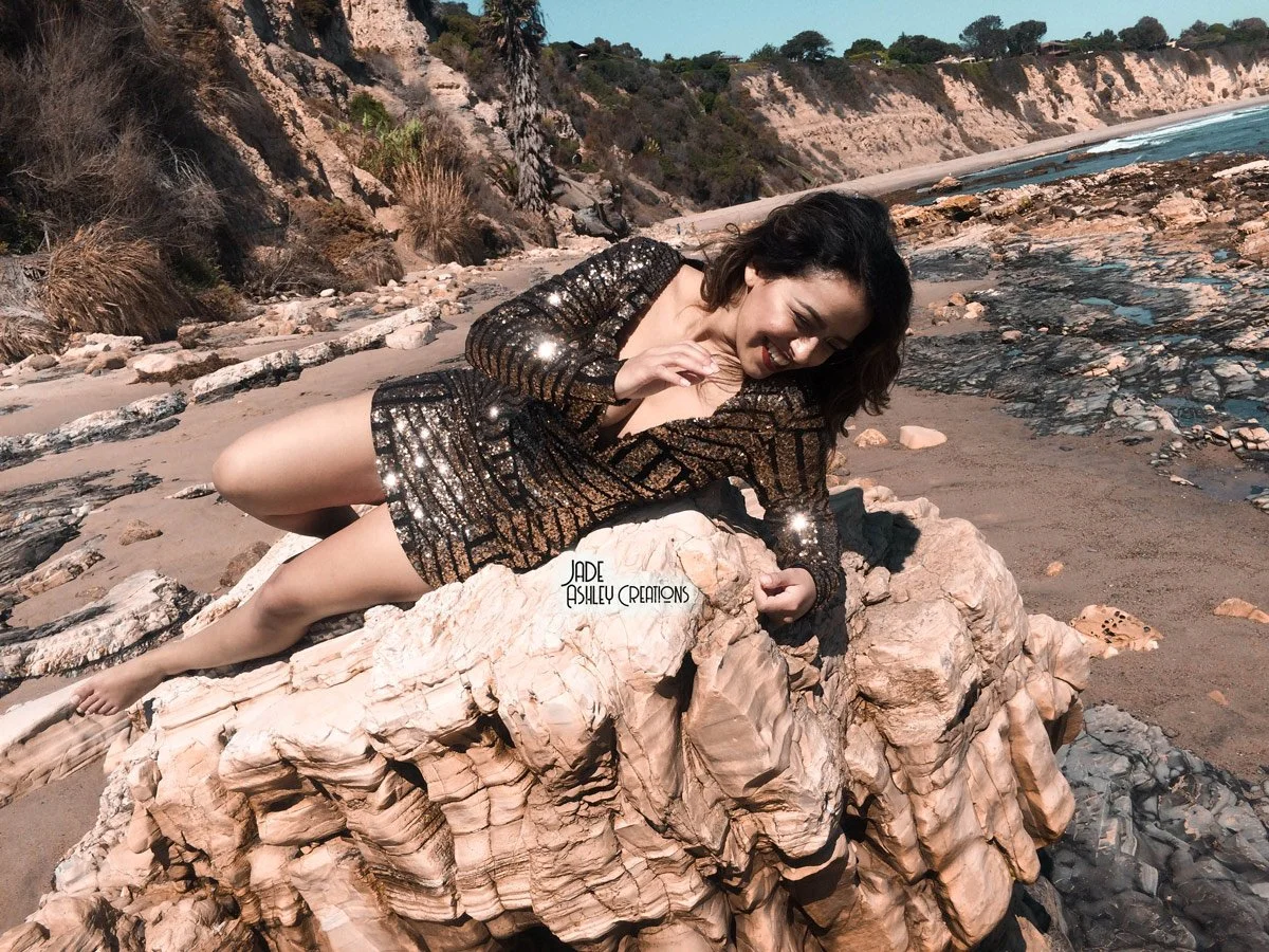 Woman in a black and gold sequined dress lying on a large rock on a beach with cliffs and ocean in the background.