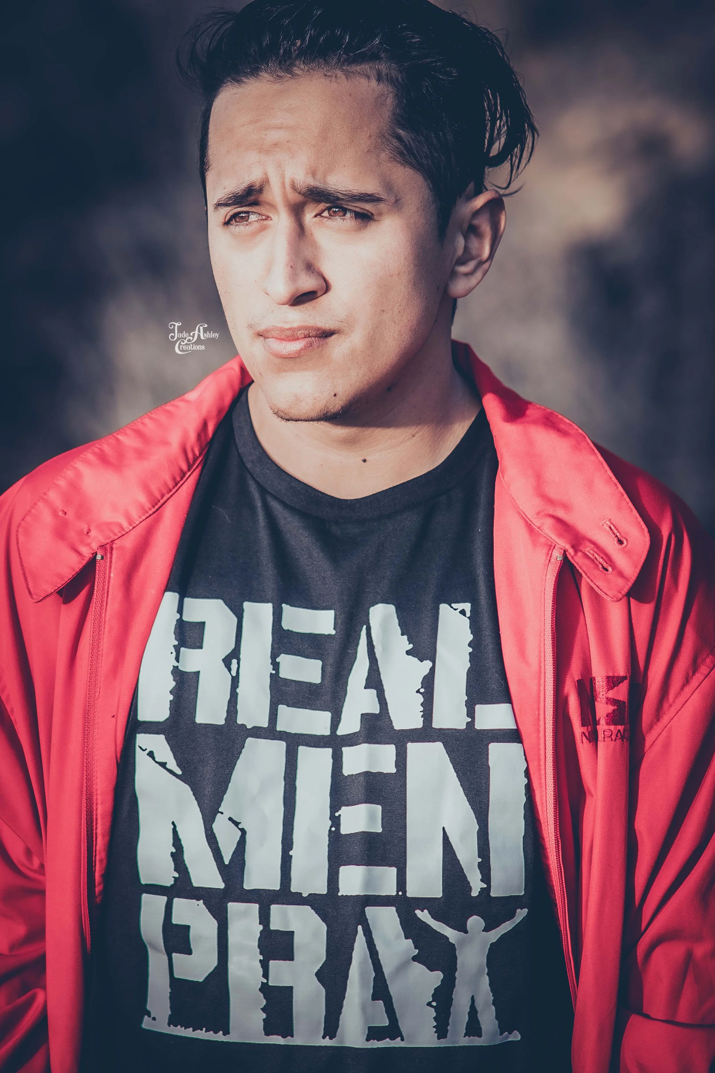 Young man with dark, styled hair looking slightly to the side, wearing a black T-shirt with white text and a red jacket, outdoors with a blurred background.
