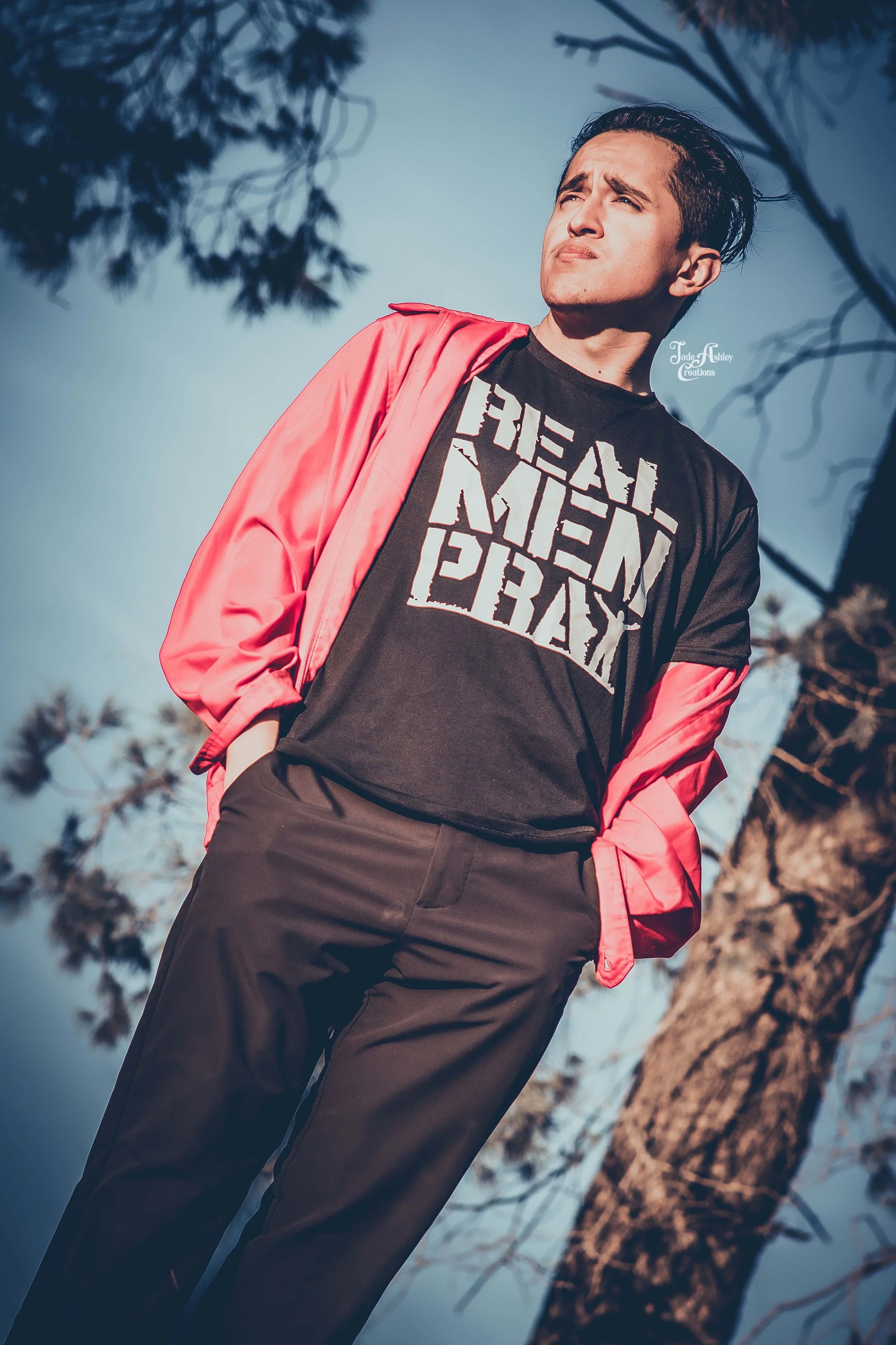A young man standing outdoors near a tree, wearing a black T-shirt with white text and a pink jacket draped over his shoulders, looking pensively into the distance.