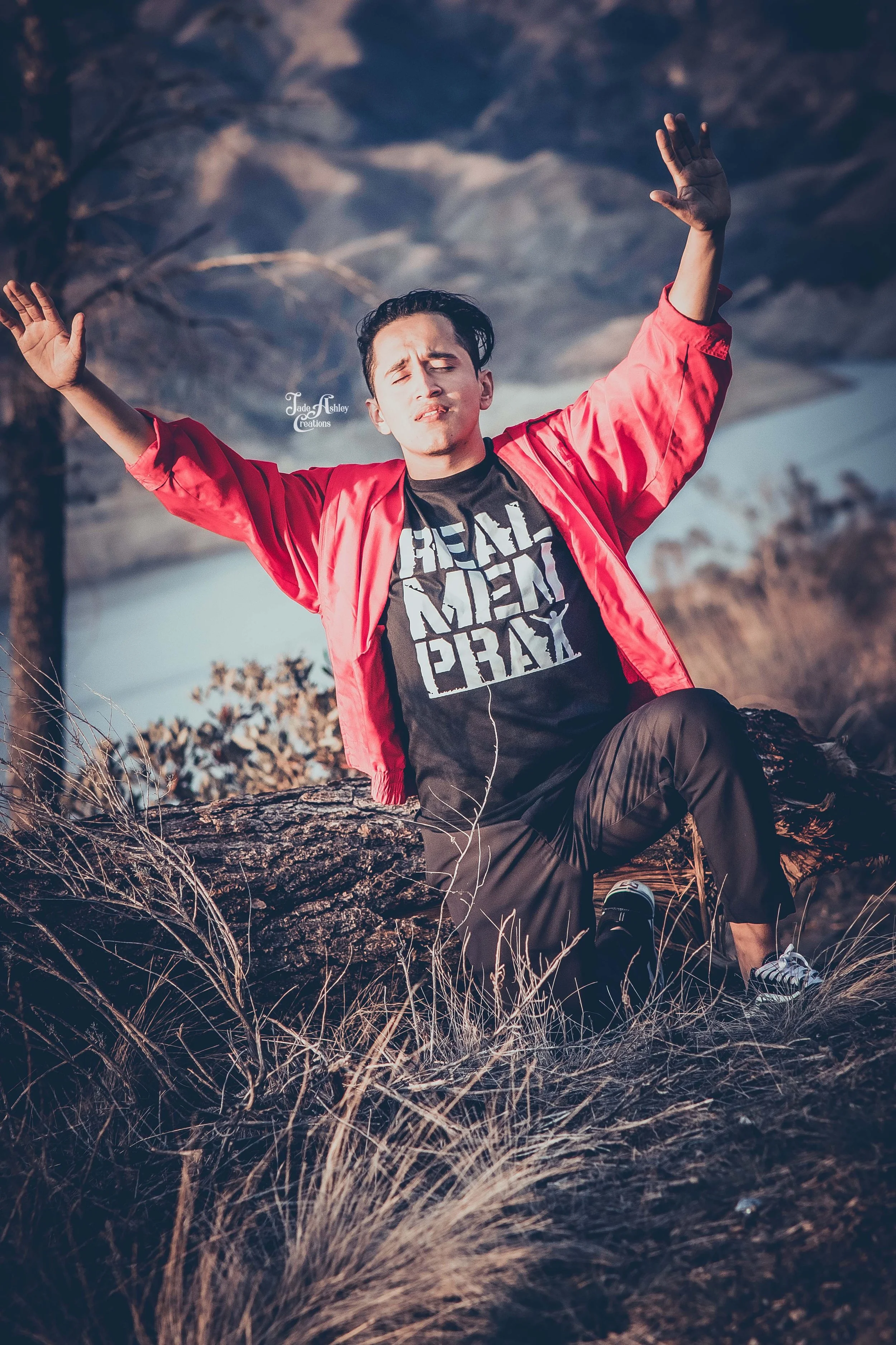 A man with dark hair in a red jacket and black t-shirt with white text, sitting on a fallen tree in a dry grassy outdoor area with trees and a cloudy sky in the background, posing with arms raised and eyes closed.
