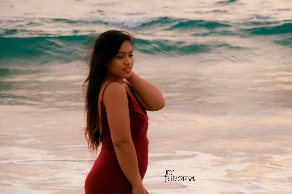 A woman in a red dress standing on the beach with ocean waves in the background, touching her neck and looking down.