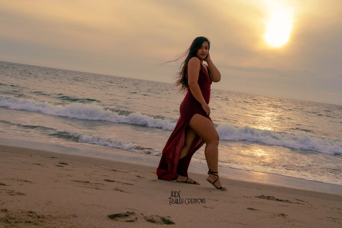 A woman in a burgundy dress with a high slit stands on a sandy beach during sunset, with the ocean waves behind her.