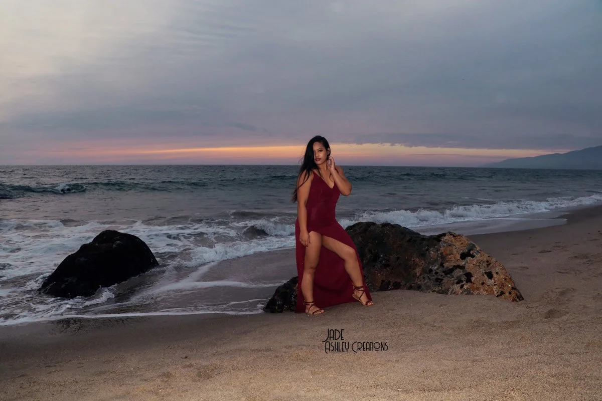 A woman in a long, burgundy dress and high-heeled sandals stands on the beach next to large rocks, with the ocean and a cloudy sunset sky in the background.