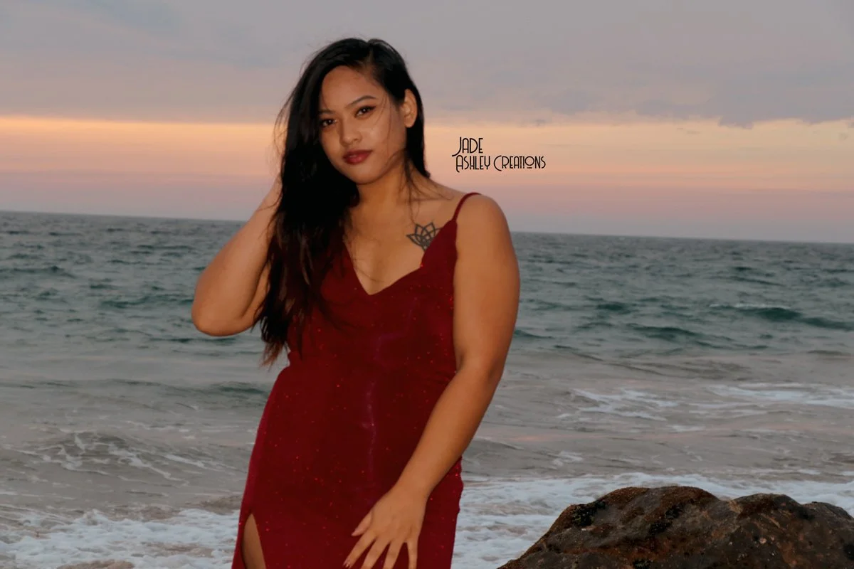 A woman with dark hair wearing a red dress standing on the beach during sunset with ocean waves in the background.