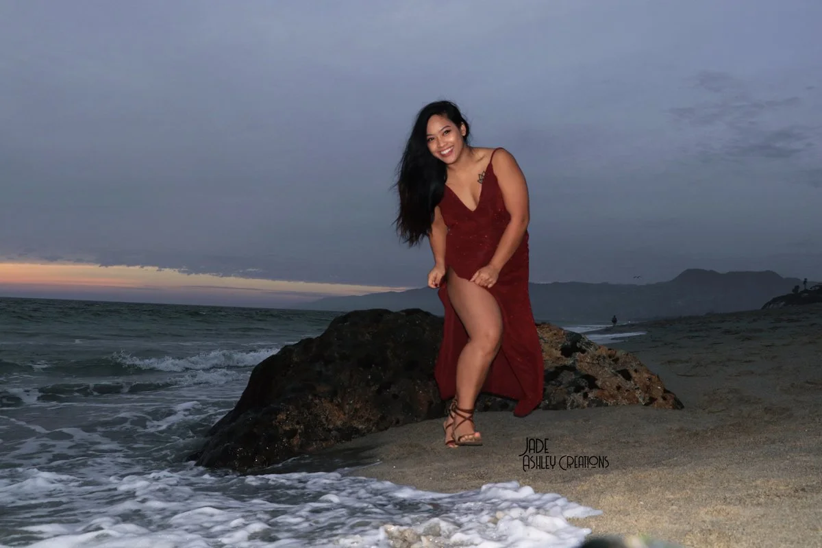 A woman in a red dress standing on rocks at the beach during sunset with the ocean waves crashing nearby.