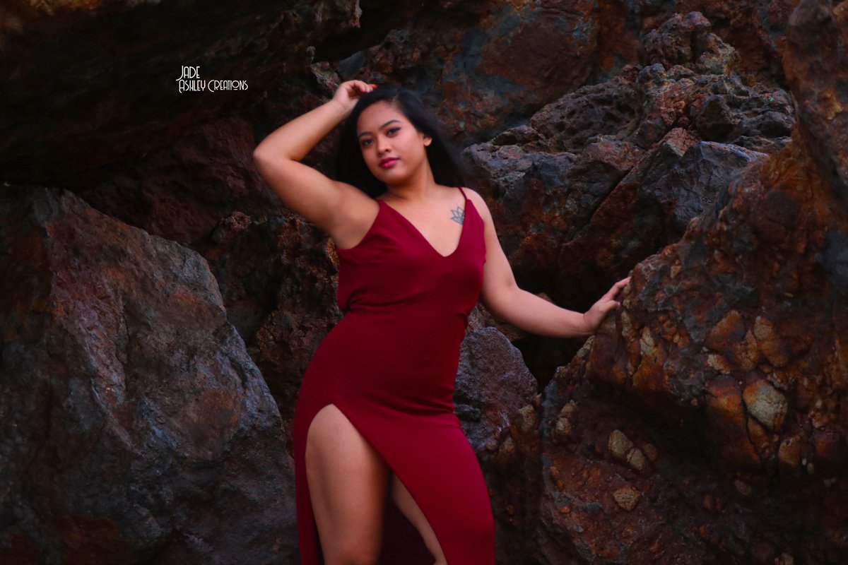 Woman in a red dress with a high slit posing among large dark rocks.