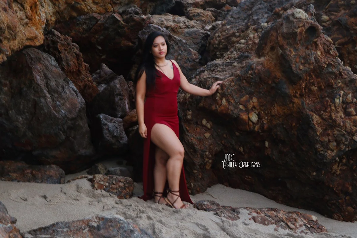 A woman in a red dress standing on sandy ground next to large rocks.